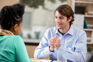 Attentive therapist listens to young adult seated at table with a smile and hands clasped 