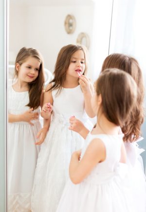 Two young girls with long hair wear white dresses and apply lip gloss in front of a mirror