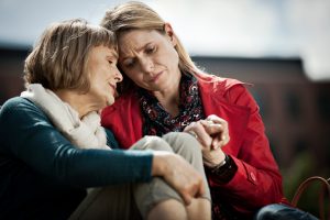 Mature woman and adult daughter sit close to each other with sad expressions on their faces. Younger woman is holding her mother's hand.
