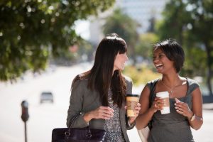 Two women in business attire walk down path holding coffee, talking and laughing