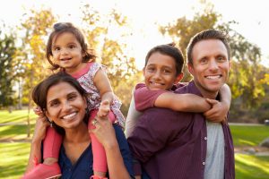 Parents of different ethnic backgrounds each hold a child on back in the park, smiling happily