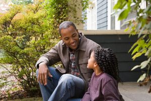 Father and daughter sit on steps outside house and have discussion smiling