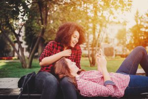 Two teenagers sit on park bench in front of sunlit trees, using a smartphone together, smiling and laughing