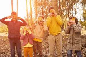 Five children of varying ages in autumn clothes pose for photo by holding leaves over their eyes