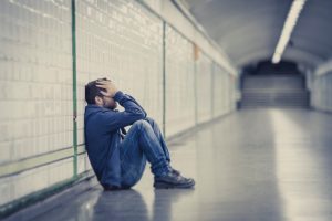 Young adult in jeans and hoodie sits against station wall, hands covering face, knees up to chest