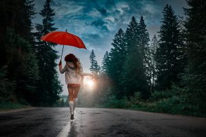 rear view of young woman jumping in spring day with her red umbrella, enjoying rainy day on a street surrounded by trees.