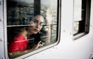 Adult with long dark hair wearing red top looks out train window with hand raised to wave goodbye