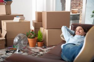 Person wearing jeans and button-down shirt lies on sofa, looking content, with open and packed boxes all over room