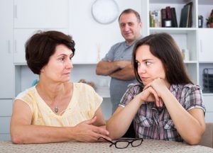 Middle-aged woman sits at kitchen table next to adult child, talking, while father stands in background with crossed arms