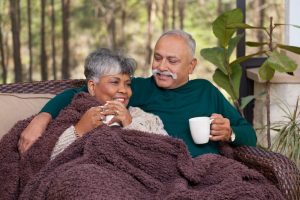 Senior couple sit on porch and talk, holding mugs, covered by a fuzzy blanket