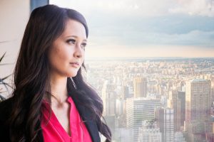 Person with long dark hair in magenta top looks out window to view of skyscrapers, apparently lost in thought