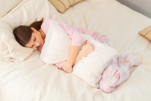 Person with long hair in ponytail wearing pink pajamas in bed, embracing a pillow with a grimace on her face