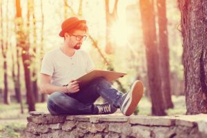 Young artist with hat and glasses sits on low rock wall drawing in large sketchbook