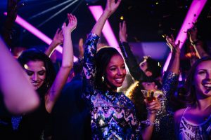 Happy young adult dancing with her friends while at a nightclub party with confetti, holding a drink on her hand