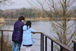Mother and daughter stand on small bridge looking over lake in winter coats. Rear shot 