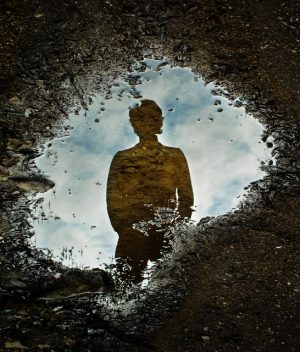 Puddle of water on ground reflects blue clouded sky and person with hands in pockets looking down into puddle