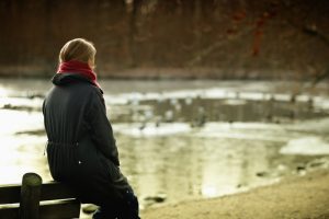 Person wearing red scarf and black coat sits on back of bench and looks out at lake