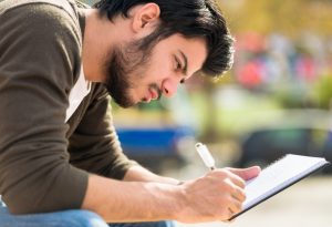 Close-up photo of young adult leaning over journal while sitting outdoors and writing 