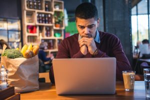 Looking at open laptop computer with worried expression with bag of groceries nearby on table