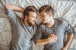 Young adult male couple lies in bed looking at each other and holding hands