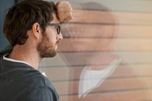 Adult with short hair, beard, and glasses leans forehead against arm on window and looks out, thinking