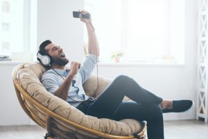 young man in headphones gesturing and keeping eyes closed while listening to music in big comfortable chair at home