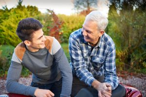Parent with white hair wearing plaid flannel shirt sits next to teenage son talking 