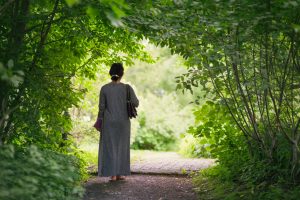 Adult in long gray wrap with hair back in clip walks through pathway under archway of green trees, standing alone, peacefully