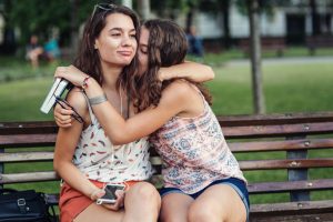 Two people sitting on park bench, one comforts other who has face in shoulder hiding