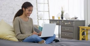 Teenager smiles while sitting on bed in decorated bedroom, legs crossed, using laptop