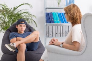 Teenager wearing shorts and backwards baseball cap sits with arms crossed, annoyed expression while counselor waits for him to speak