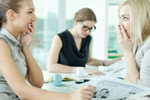 Two colleagues sitting at office table laugh and whisper about third colleague working nearby