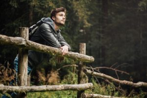 Young adult hiker with backpack in the forest leans on a wooden fence looking lost in thought