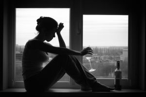 Black-and-white photo of person sitting in window with glass and bottle of wine, hand pressed to head