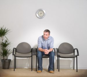 Bored-looking person sits alone in waiting room, leaning forward, hands folded