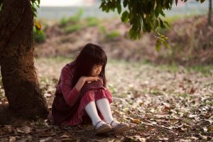 Distressed child with long dark hair sits under tree, knees drawn up to chest and hands on knees