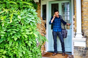 young adult in plaid shirt and jeans with leather bag speaks on phone while leaving house