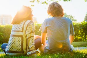 Rear view of parent and teen sitting on grass, sharing music through headphones on a sunny day