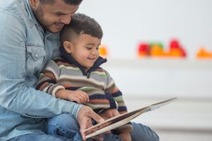 Young child sits in lap of parent with short hair who reads large picture book. Both parent and child are smiling