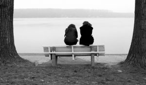 Photo shows rear view of two people with long hair wearing coats sitting on back of bench by water