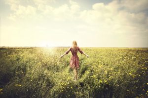 Person with long blonde hair walks through lush green field with arms outstretched