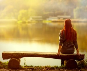 Rear view of person with long red hair sitting on bench at sunset looking over lake