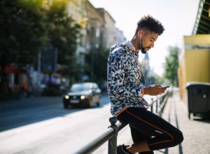 Side view of young adult with short hair in athletic clothes half-seated on railing wearing headphones and using phone