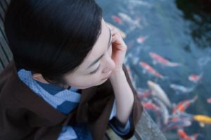 Resting chin in hands on side of low footbridge, looking down into koi pond thoughtfully