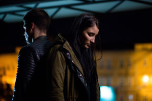 Couple dressed in light jackets stand outside at night at distance from blurred lit building, facing away from each other, looking down