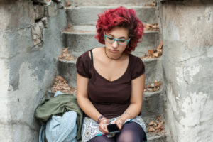 Person with short curly red hair, blue glasses, wearing tights and skirt sits on leaf-strewn stairway and reads on phone with somber expression