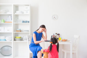 Parent with long ponytail and blue dress sits at desk, forehead supported with hand, as child with pigtails approaches with toy