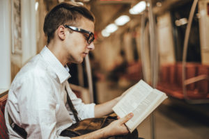Young adult professional with short hair and glasses reads book on train, peacefully making way through commute