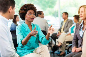 Black person with aqua blouse speaks at seminar on diversity to group of seated individuals