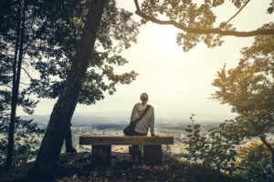 Rear view of person wearing shoulder bag and hoodie with blonde ponytail sits on bench on top of the hill looking down on town